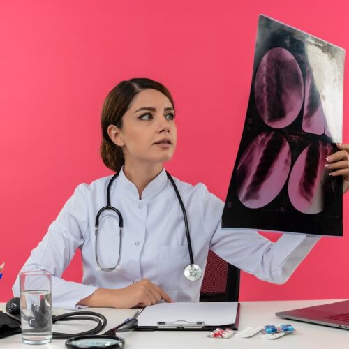 pleased-young-female-doctor-wearing-medical-robe-with-stethoscope-sitting-desk-work-computer-with-medical-tools-holding-looking-x-ray-with-copy-space_141793-37718